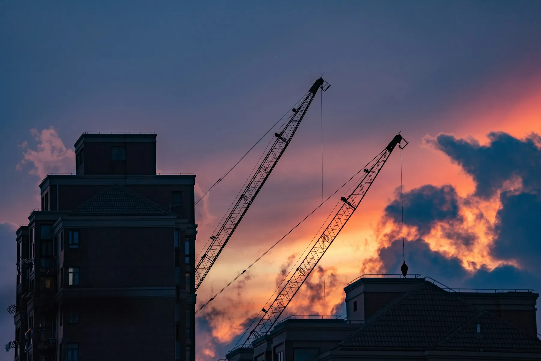 Silhouettes de grues de chantier se découpant sur un ciel de coucher de soleil, à côté d’immeubles résidentiels